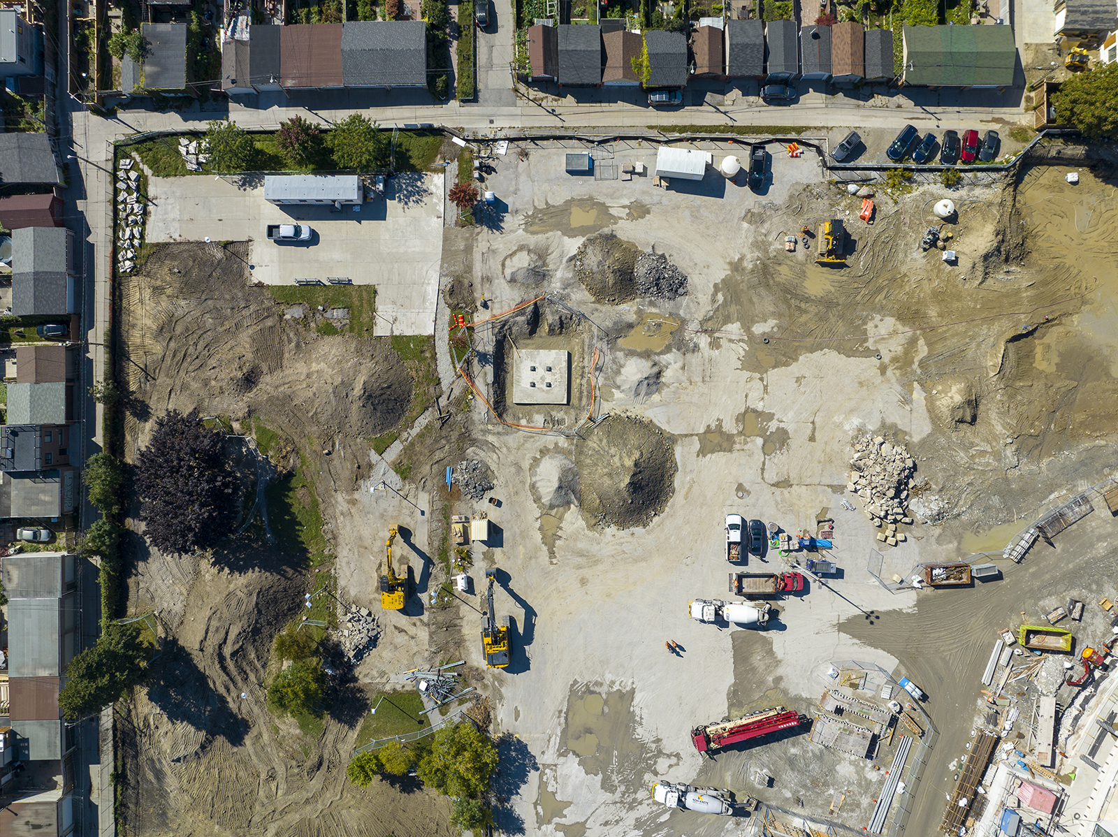 An aerial photo taken from above by a drone of the construction site at Wallace Emerson Park. The image shows construction equipment, cars and trucks at the project site, with small flat areas that have been excavated in preparation for below ground construction. Residential homes line the perimeter of the construction site. 