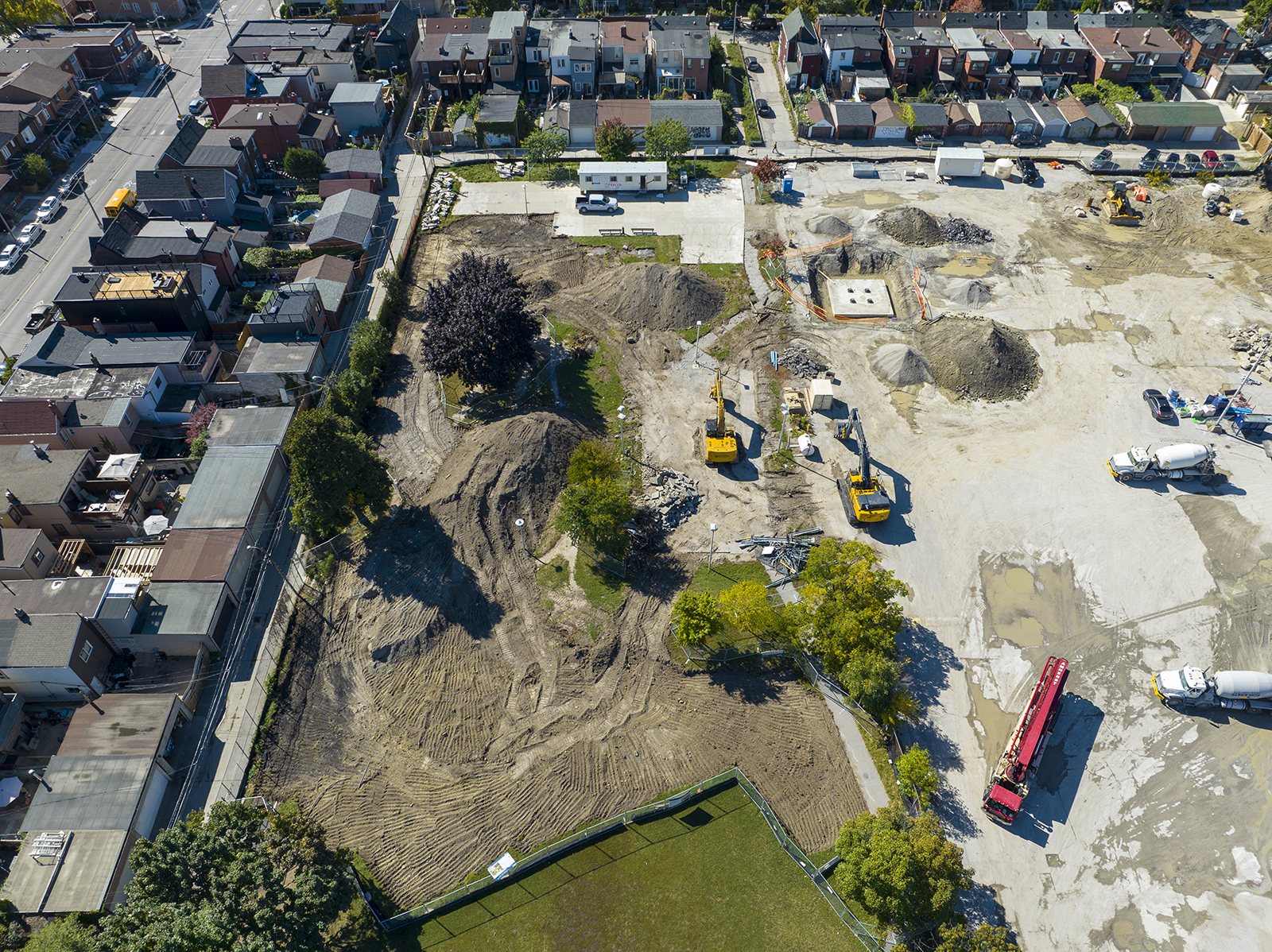 An aerial photo facing east taken by a drone of the construction site at Wallace Emerson Park which shows the excavation occurring parallel to Dupont Street. Large mounds of dirt and yellow cranes are shown and the surrounding residential homes which border the fenced off construction site. 