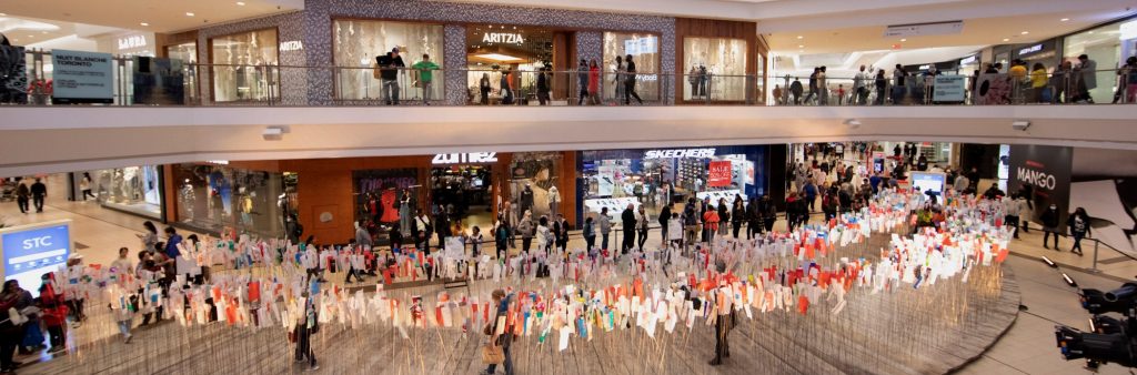 Art installation in Scarborough Town Centre of ties on posts at shoulder height. People walk through installation and other people look on from the mezzanine above.