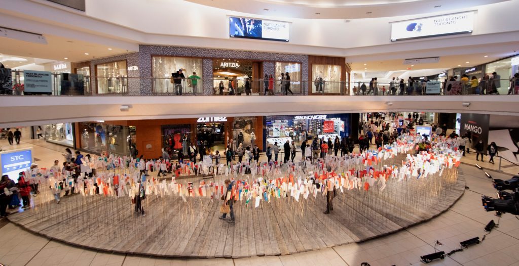 Art installation in Scarborough Town Centre of ties on posts at shoulder height. People walk through installation and other people look on from the mezzanine above.