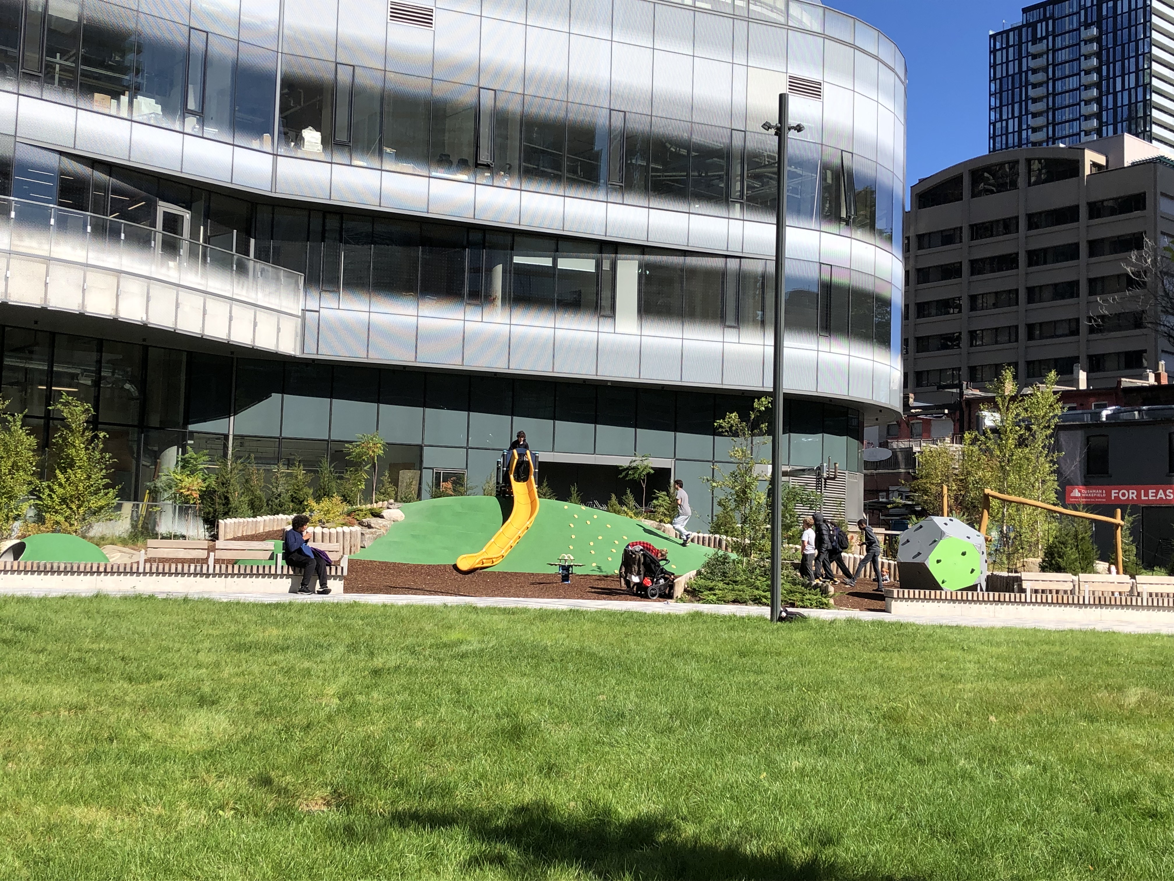 A photograph of Dr. Lillian McGregor Park which shows the playground and children playing on a yellow slide and climbing features. An open lawn area is in the foreground.