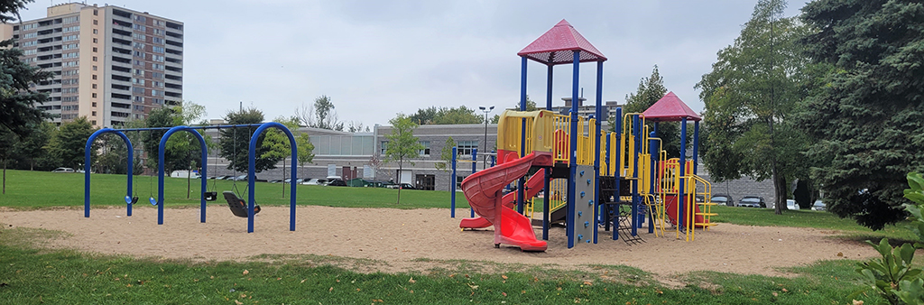 A photograph of the playground in Scarborough Village Park showing a large playground structure in red, blue and yellow and a swing set on top of sand. Open lawn space surround the playground area.