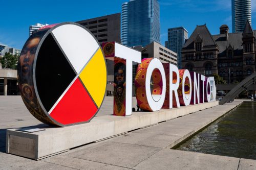 Colour photograph of a large exterior sign spelling the word Toronto with a medicine wheel and Toronto's old City Hall in the background.