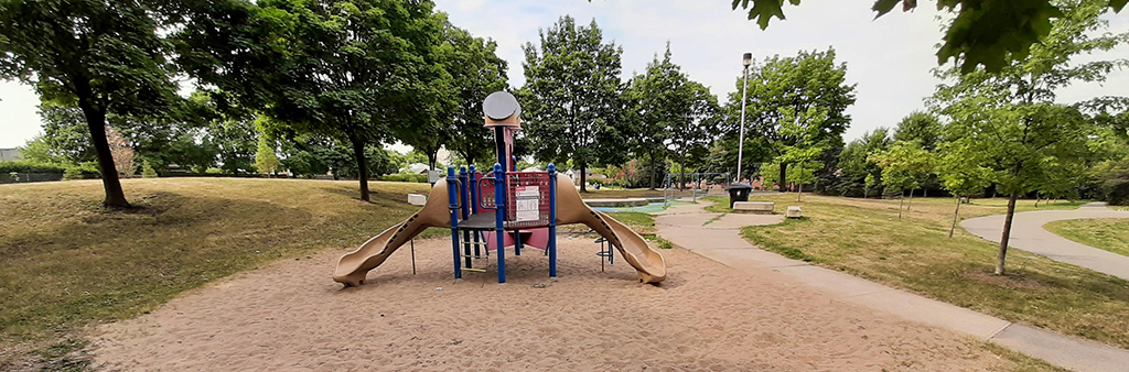 A photograph of Heathrow Park Playground which shows a small play structure in the foreground on top of sand. The playground is surrounded by grass, winding pathways and mature trees.