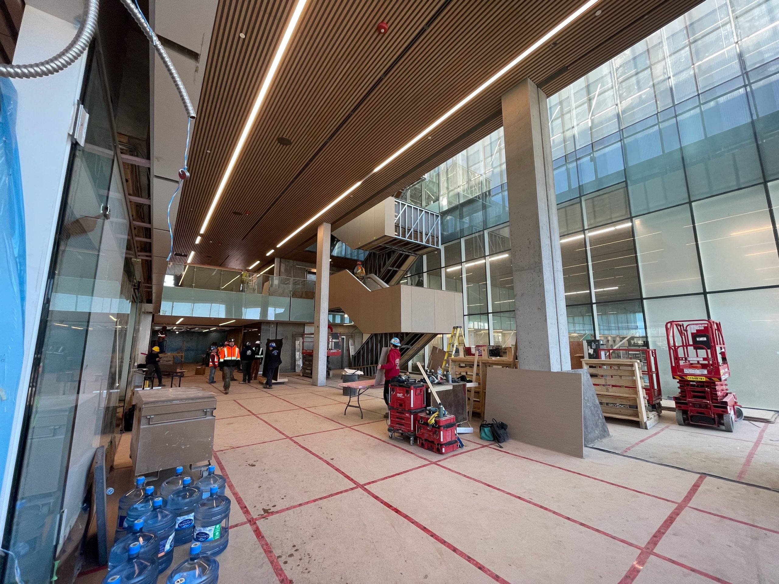 A photograph showing the construction progress of the main entrance of the new Ethennonnhawahstihnen’ Community Recreation Centre. The photo shows the main level and second level, connected by a large staircase in the centre, parallel to a floor to ceiling wall with glass panels. 