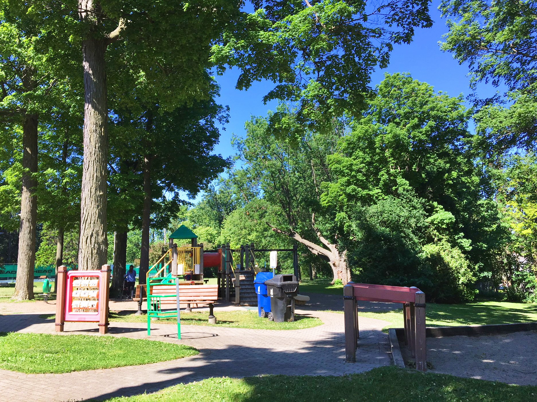 A photograph of Banbury Park Playground before improvements, which shows various stand-alone play features around the playground area, with a medium sized play structure in the background. The playground is surrounded by mature trees and various pathways.