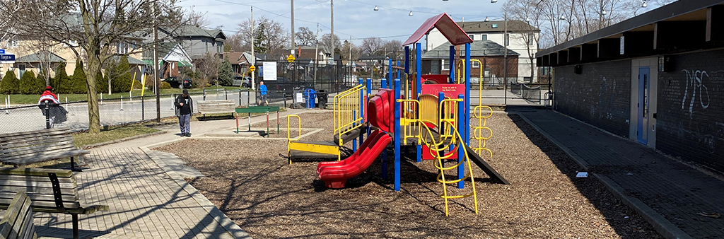 A photograph showing the playground area and adjacent clubhouse building in Topham Park. The playground equipment is shown in red, yellow, and blue and is surrounded by a pathway with two benches.
