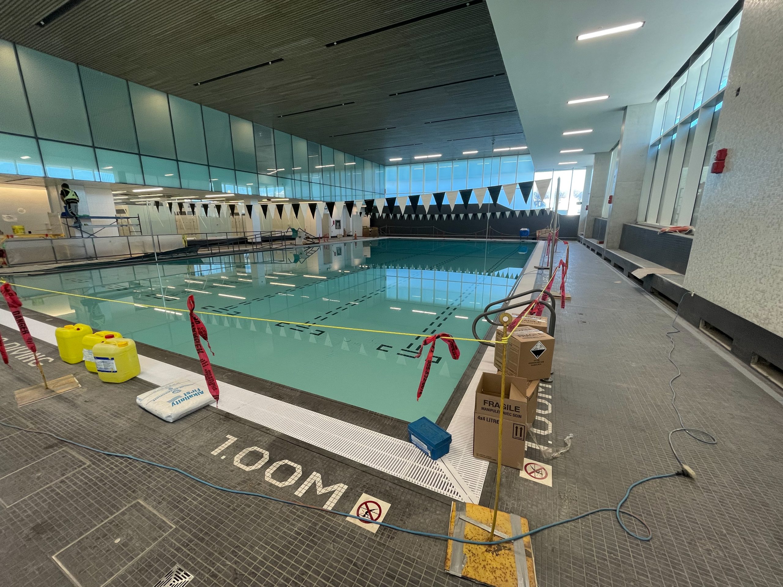 A photograph showing the construction progress of the indoor swimming pool in the new Ethennonnhawahstihnen’ Community Recreation Centre. The photo shows a substantially completed swimming pool, with a pool deck and frosted glass panel design features. 