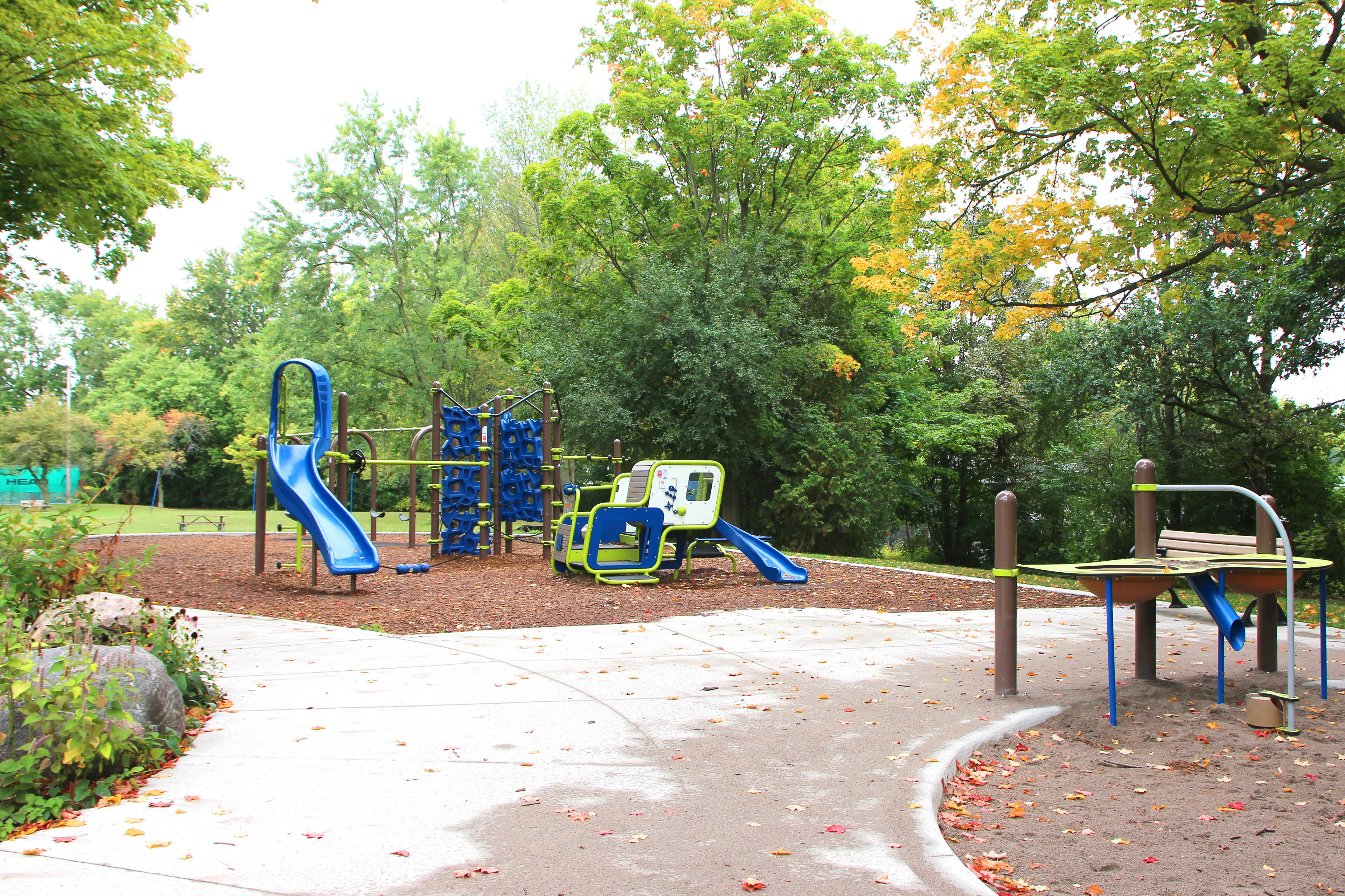 Banbury Park Playground after construction showing a circular seating area with stones in the foreground, an accessible sand table in blue and lime green parallel to two benches, and a play structure in the background with a blue slide and climbing features in blue and green.