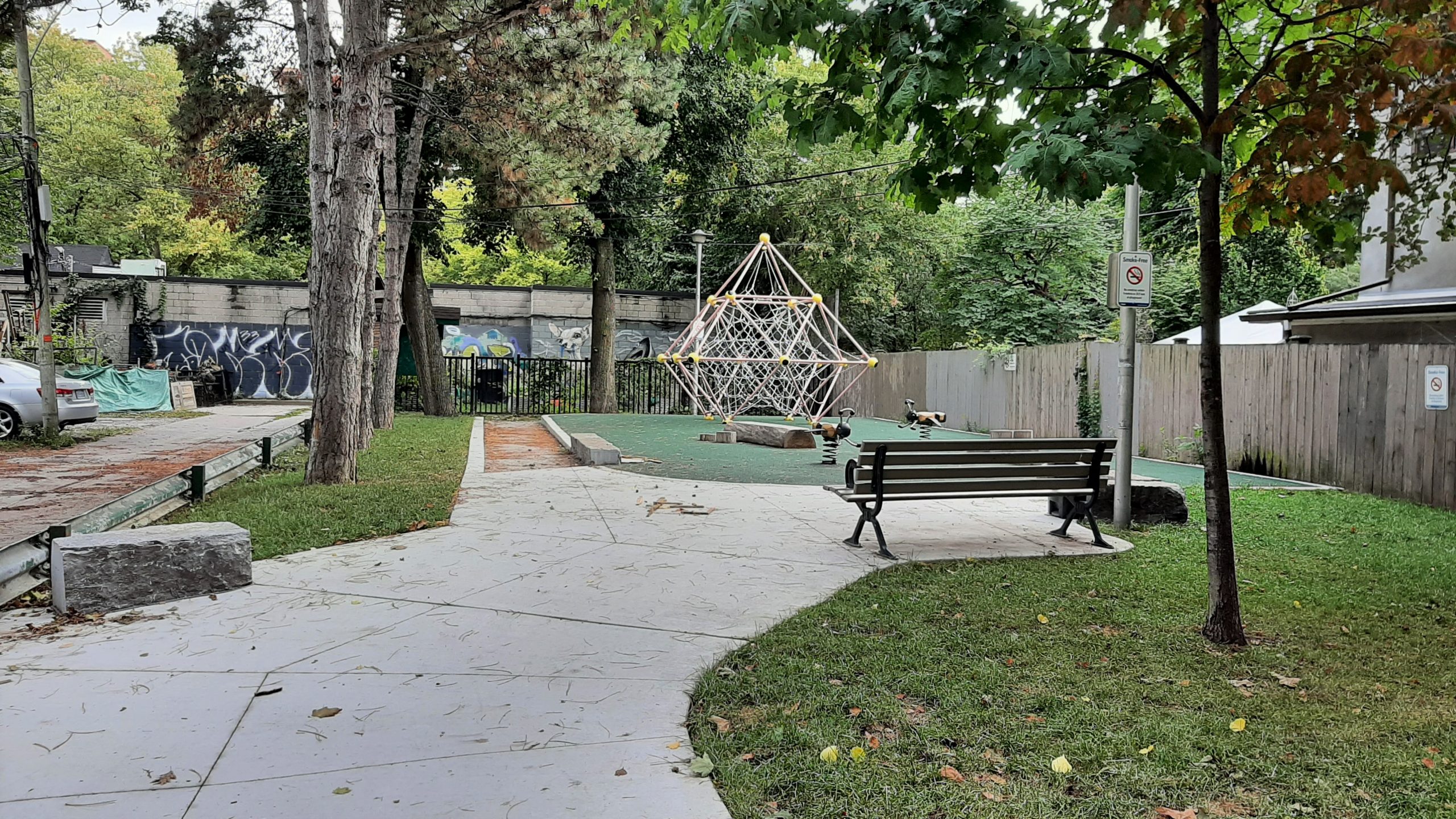 A photograph of Melbourne Avenue Parkette Playground after improvements, which shows a wide concrete path leading to the small playground area, with a bench and large geometric climbing structure in the centre. The playground also features two spring toys, a balance log, and green rubber surfacing.