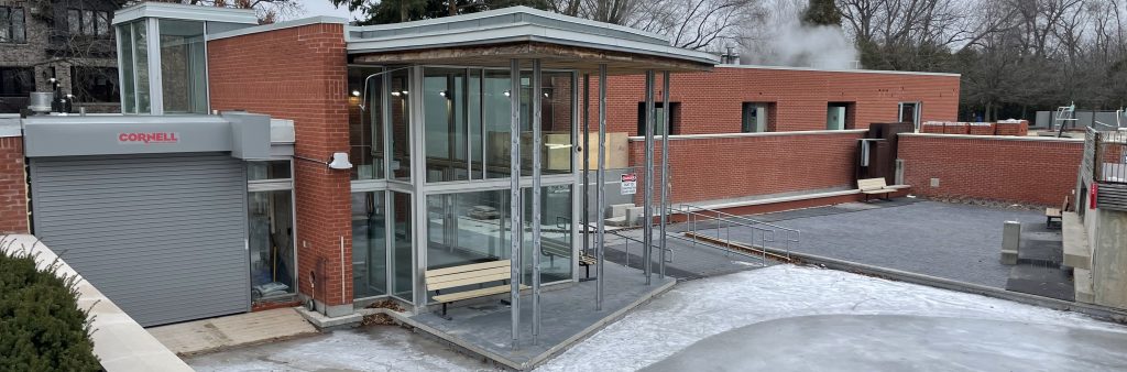 A photograph of the exterior of Ledbury Park Building after construction showing the small ice rink in the foreground and brick building with tall glass windows in the background. Attached to the building is a small garage with a grey door. A bench on a concrete pad outside the building is facing the ice rink.