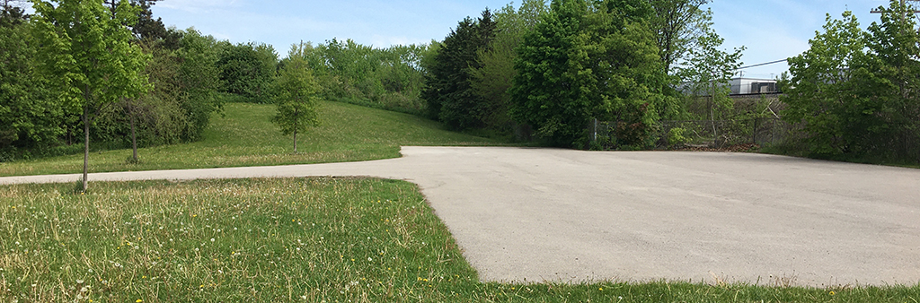 A photograph of North Park, looking north towards the parking pad. The photo shows an asphalt path leading to a square parking pad surrounded by an open lawn.