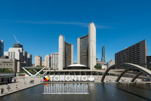 Large, outdoor square in summer with reflecting pool in foreground and curved building towers in background.