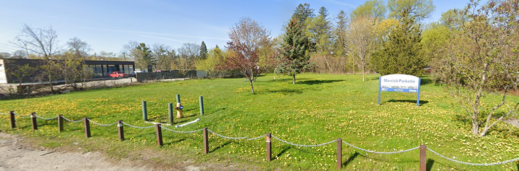 A photograph of the existing Morrish Parkette, taken from Morrish Road. The park is a large expanse of grass, with some scattered tree plantings and many tree plantings to the rear of the park. There is a post and chain fence around the front of the park, as well as a blue and white park sign.