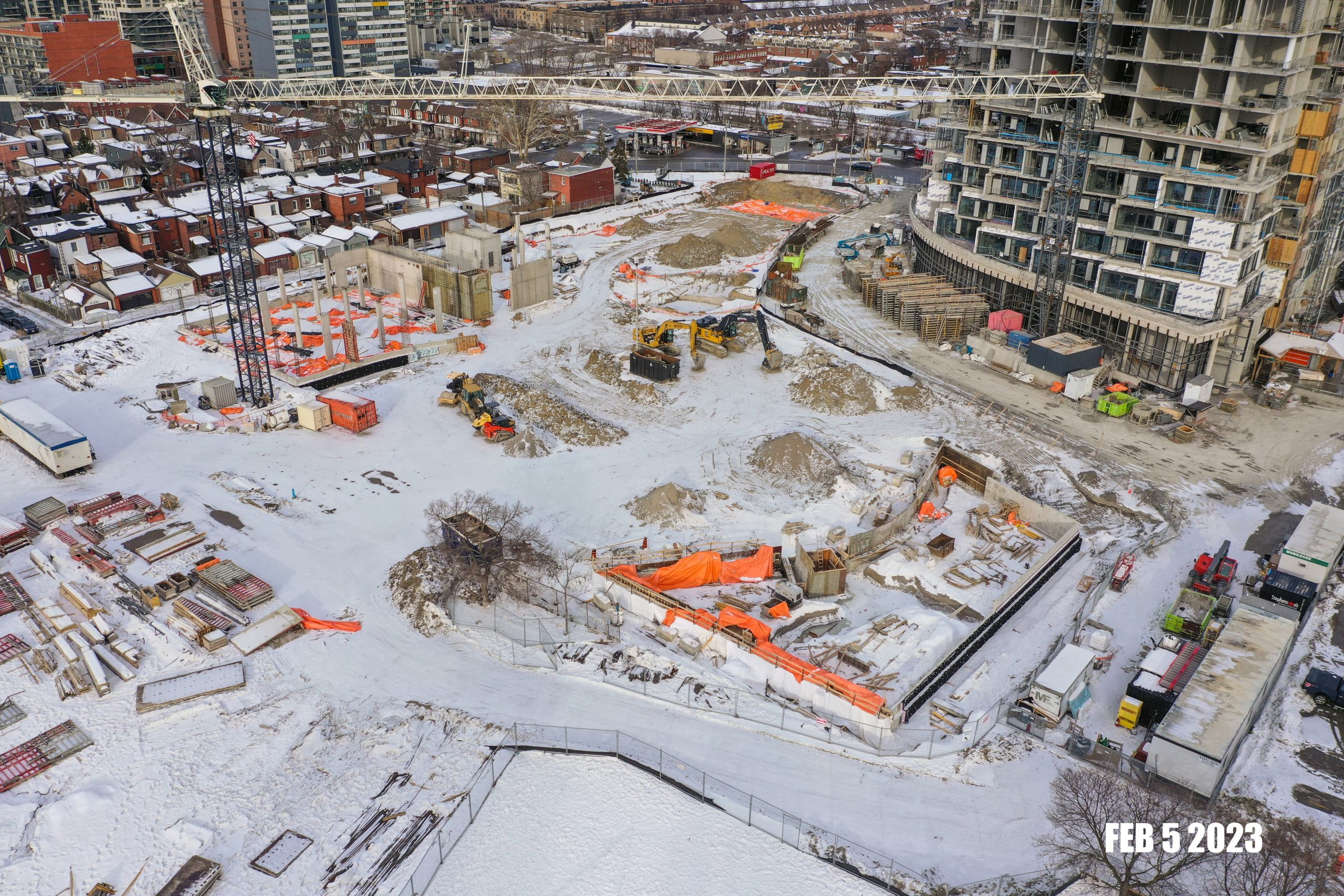 An aerial photograph of the construction site at Wallace Emersion Community Centre on in February 5, 2023, which shows a large crane and multiple excavators and construction equipment. The construction area is predominantly flat, with soil and sand throughout. A thin layer of snow covers the construction area and surrounding residential homes. 