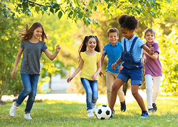 Five children playing soccer in a park on grass with trees in the background