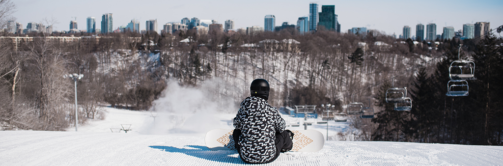 A person wearing a white and black ski jacket and snow pants, sitting at the top of a ski hill with a snowboard. Off into the distance is the ski hill, trees and a city landscape.
