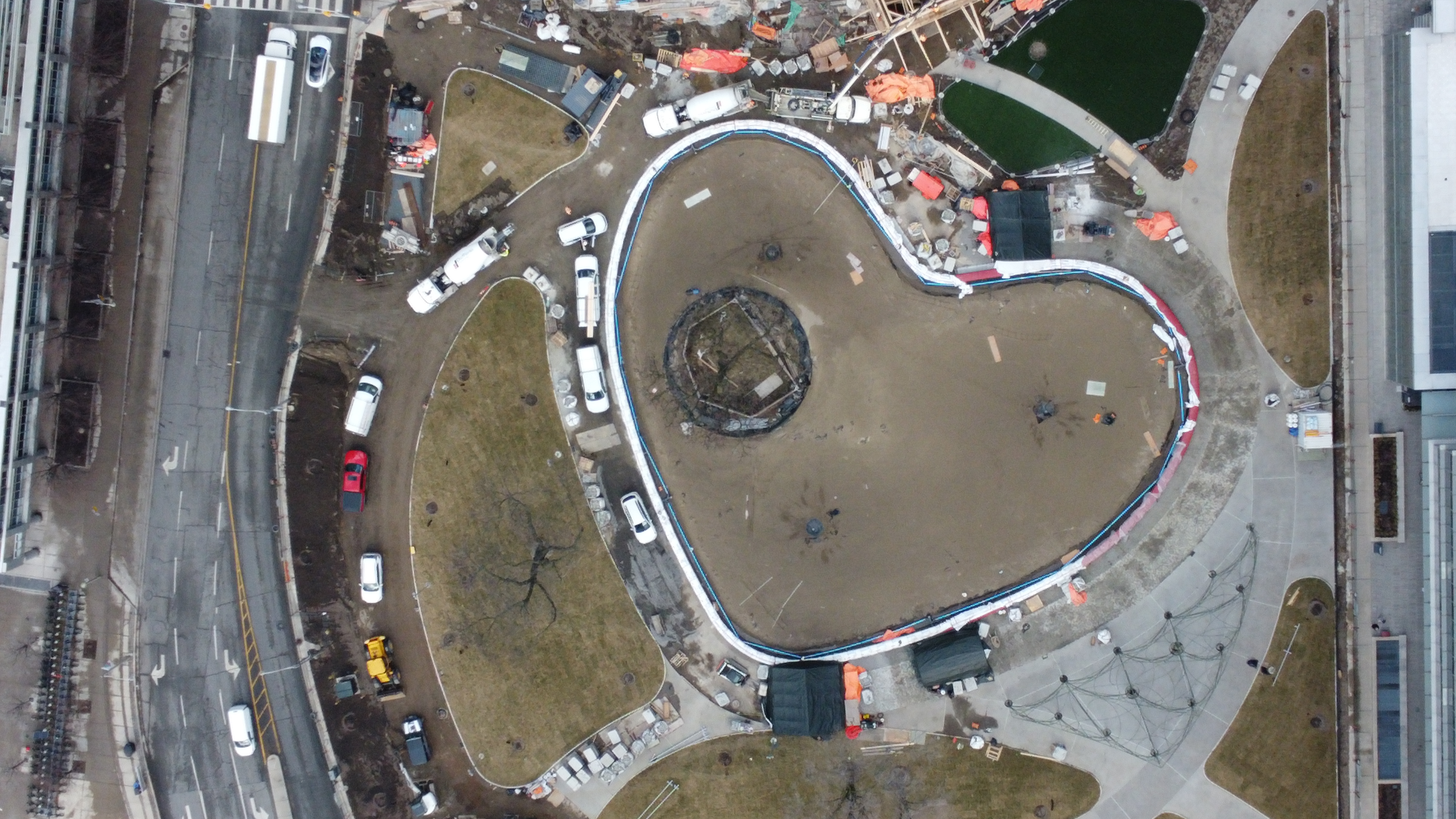 An aerial image taken by a drone showing the construction progress for the new park, which shows the centre of the park outlined as a large heart shape and ongoing landscaping around this area, including dirt paths with patches of grass in irregular rounded shapes. The park is a construction site, with construction equipment ,cars, and dirt. 