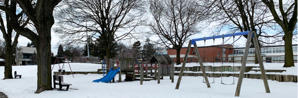 A photograph of Dunlace Park Playground taken during the winter with snow on the ground which shows the swing set with two swings in the foreground and a wood play structure in the background. The playground are is surrounded by mature trees.