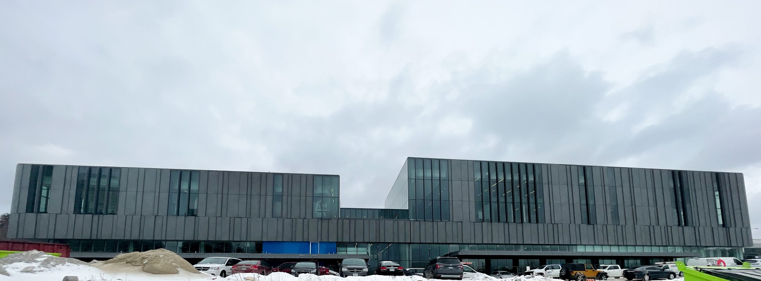 A photograph of the exterior of the new Ethennonnhawahstihnen' Community Recreation Centre & Library which shows a long rectangular multi-floor building with tall glass window panels and dark grey panels. 