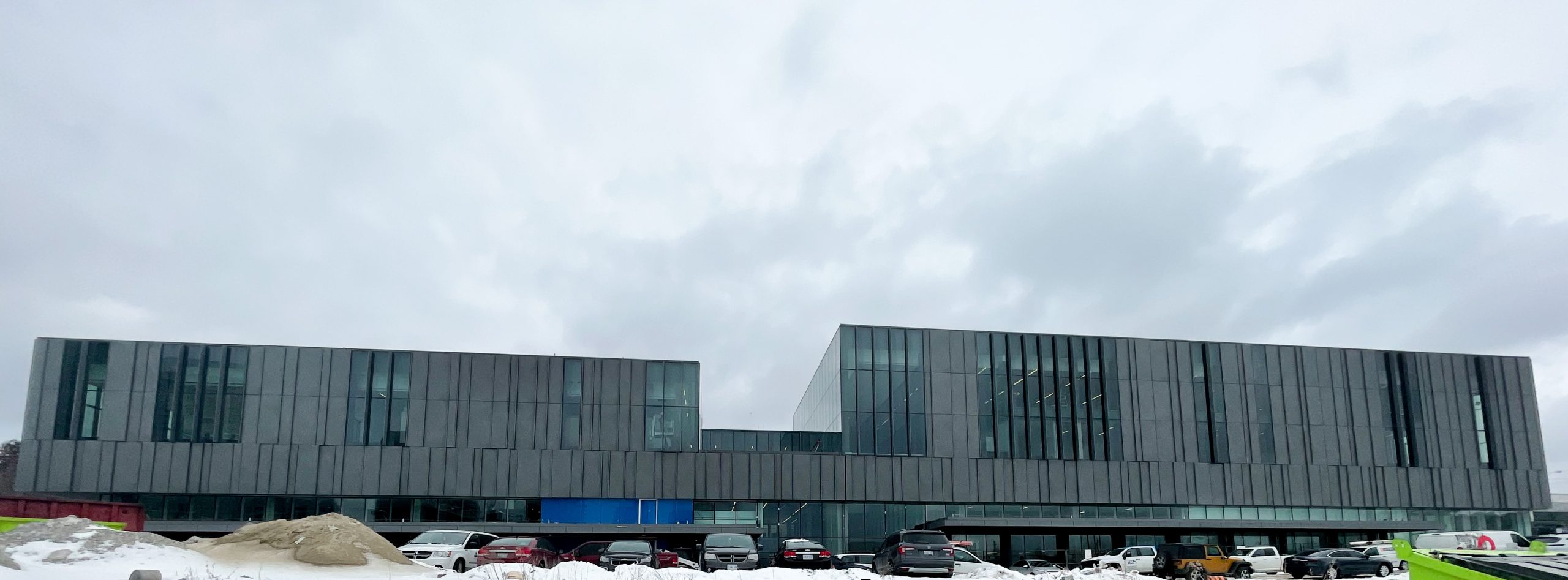 A photograph of the exterior of the new Ethennonnhawahstihnen' Community Recreation Centre & Library which shows a long rectangular multi-floor building with tall glass window panels and dark grey panels. 