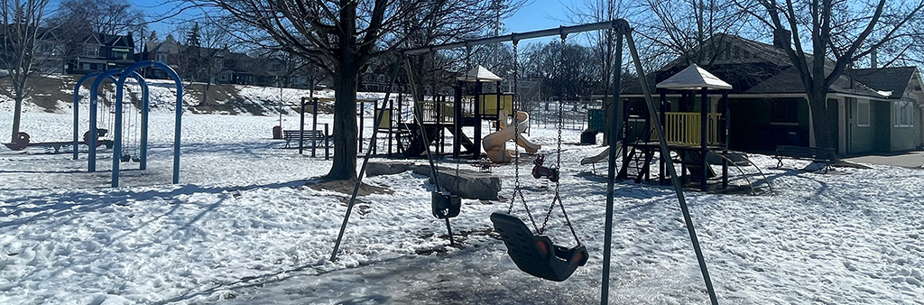 A photograph of Greenwood Park Playground taken during the winter with snow on the ground. A swing set is shown in the foreground and a junior and senior play structure is in the background.