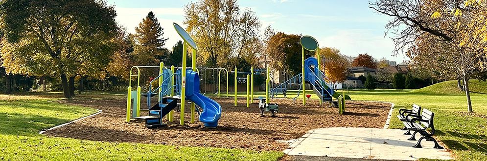 The playground after construction, featuring a mulch surface, a junior play structure in the foreground, a senior play structure, a swing set with three bays, play panels, and springer toys throughout. Additionally, a concrete seating area with two benches is located on the east side of the playground.