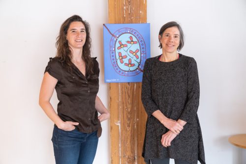 Two people stand facing the camera and between them an artwork attached to a vertical wooden beam