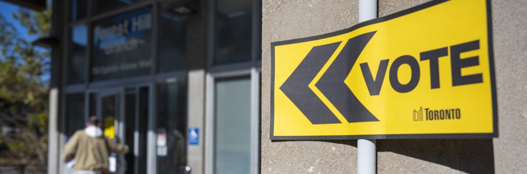 Toronto Elections yellow vote sign on a door directing to a voting location, with person blurred in the background going into the building.