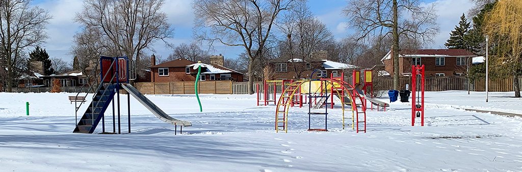 A photograph of Stephen Leacock Playground taken during the winter with snow on the ground. The photo shows a small stand-alone slide and stand-alone domed climber in the foreground and a play structure and swings in the background.