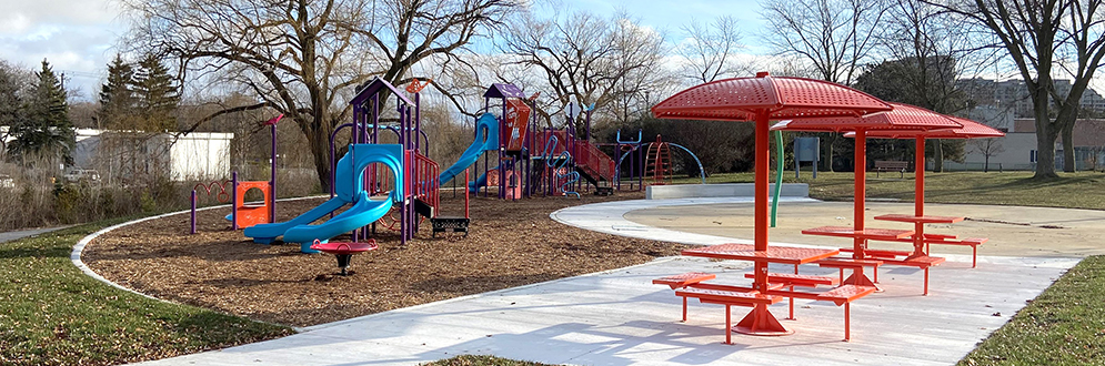 The new Stephen Leacock Park playground looking east from the new walkway. The playground is surrounded by a walkway with covered picnic tables and a splash pad is in the background.