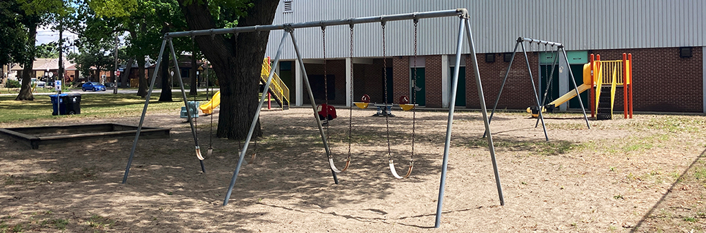 Runnymede Park eastern playground as viewed from the west. There are two sets of swings and a large shade tree in the foreground. The George Bell arena and two slides are in the background.