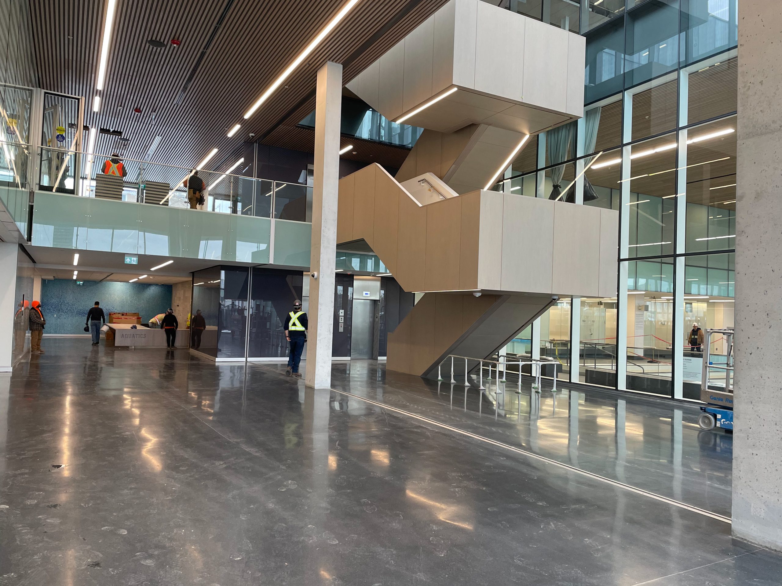 A photograph of the interior lobby area of the new Ethennonnhawahstihnen' Community Recreation Centre & Library while under construction, which shows a large feature staircase that connect the main floor and second floor. The second floor overlooks the main floor. To the left of the main level is a floor to ceiling glass wall into the swimming pool area. 