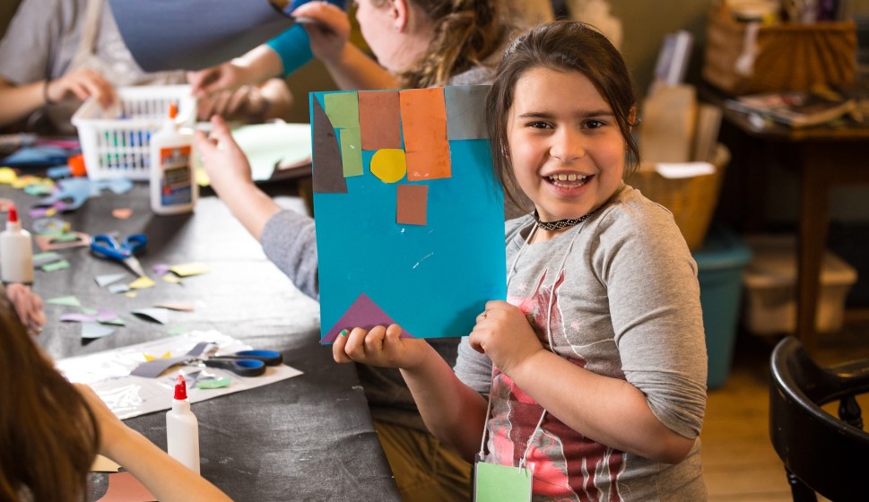Child smiles and holds up their handmade artwork. People working on art at a table in the background.