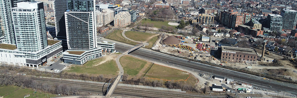 Bird's eye-view photograph of 10 Ordnance Street and 801 Wellington Street West, as well as surrounding context. The photograph is taken from above Garrison Common and faces north west. The Garrison Crossing pedestrian and cycling bridges are seen in the photo, as well as the linear open space system heading north towards Trinity Bellwoods Park.