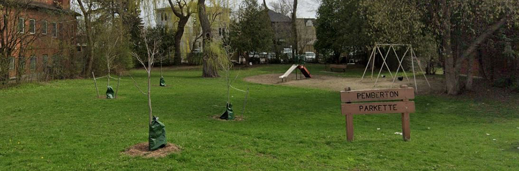 A photograph of Pemberton Parkette which shows the wood park sign in the foreground and a small playground in the background, surrounded by young and mature trees and grass.