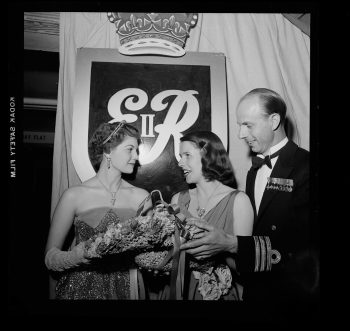 Photograph depicts two women in ball gowns and one naval officer wearing formal mess dress uniform