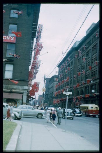 Photograph depicts store fronts decorated with Union Flags and Canadian Red Ensigns