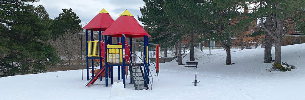 A photograph of Scarden Park Playground taken during the winter which shows a bench facing the playground area in the foreground and one play structure in the background. Snow covers the ground and mature trees surround the area.