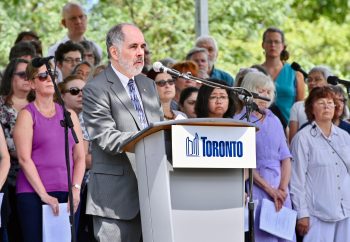 Toronto Poet Laureate Al Moritz speaks into microphone behind podium with crowd in background