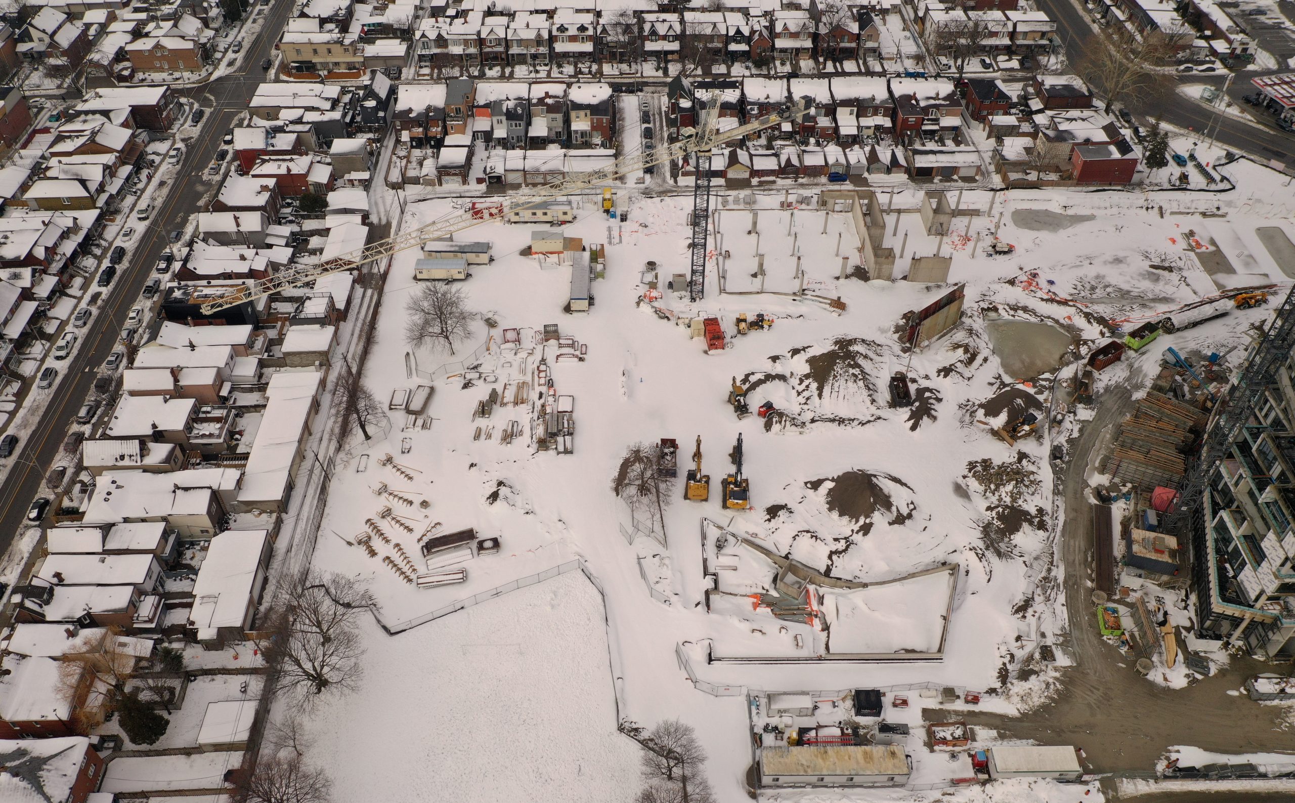 An aerial photograph of the construction site at Wallace Emersion Community Centre on March 4, 2023, which shows a large crane and multiple excavators and construction equipment. A thin layer of snow covers the construction area and surrounding residential homes.