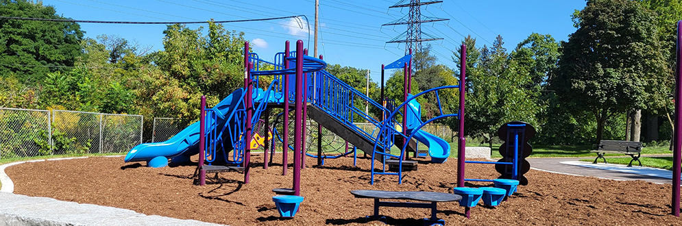 Photo of the new Lambton Park playground looking northeast. There is armourstone seating, a stand-alone climber, and a senior play structure with a triple slide in the foreground, and a junior play structure with a curved slide visible in the background.