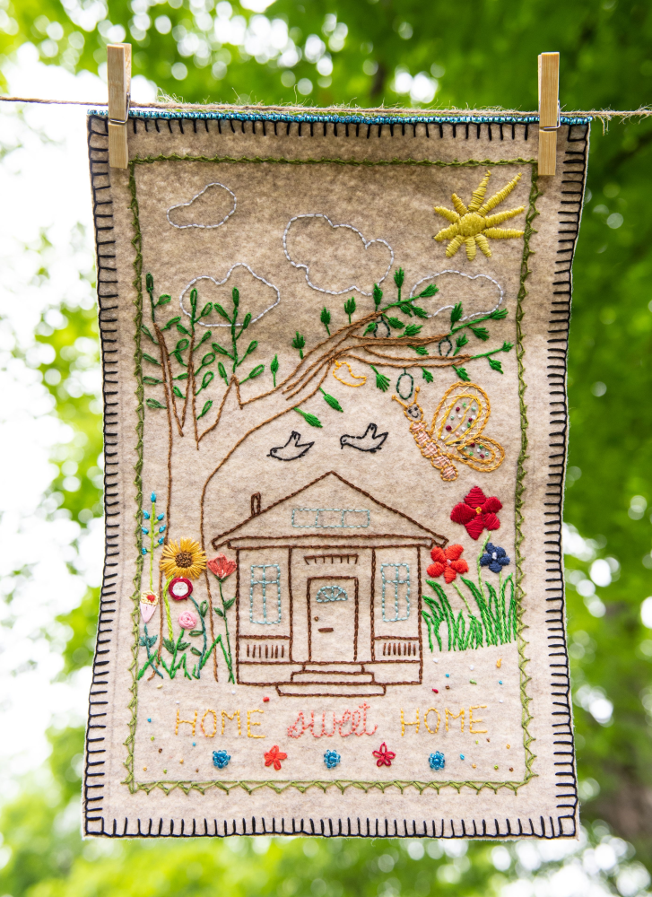 Photograph of an embroidered fabric held onto a string with clothing pegs with trees in background. Embroidery of sun, tree, butterfly, birds, flowers, a house and the words home sweet home