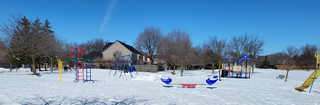 A photo of Elmbrook Park Playground taken during the winter when there is snow on the ground showing a see-saw and stand-alone slide in the foreground, and various play structures in the background. The playground is surrounded by open space and mature trees.