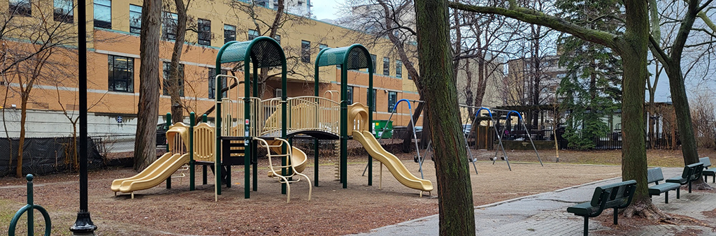 Photograph of Tichester Park Playground on a rainy day taken from the sidewalk adjacent to the playground showing a playground structure in yellow and green and a stand-alone swing set next to it. Benches face the playground area.