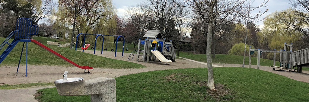 A photo of Wigmore Park Playground which shows a drinking fountain and stand-alone straight slide in the foreground and a playground in the background. A pathway connects to the play area, which is surrounded by mature trees and grass.
