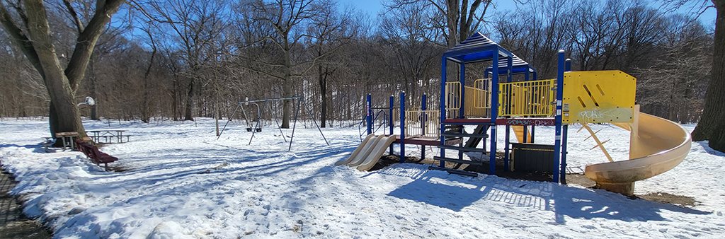 A photo of Magwood Park Playground taken during the winter which shows a combined playground structure with slides and a swing set in the background. The playground is surrounded by mature trees and grass covered in snow.