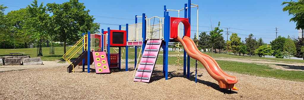 A photograph of North Kipling Park Playground which focuses on the large play structure with various play components in colours like red, yellow and blue. A large field with grass and tress is in the background.