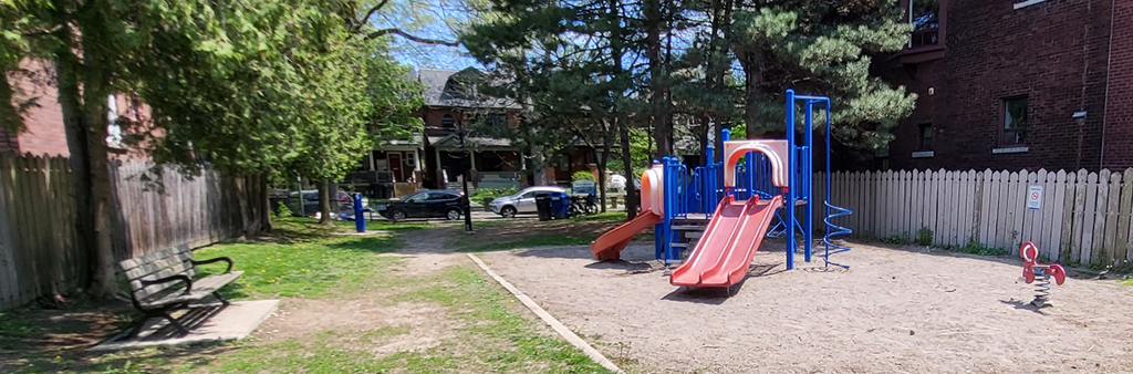 Photograph of Beaty Avenue Parkette Playground taken in the spring which shows a small play structure in blue and red on top of sand. The playground is between to residential homes.