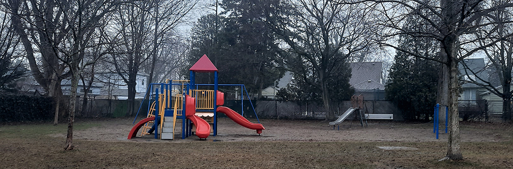 A photograph of Delma Park Playground taken from a distance on a rainy wet day showing the entire playground area. In the centre of the photo, is the combined junior/senior play structure with three red slides. The playground is on top of sand.
